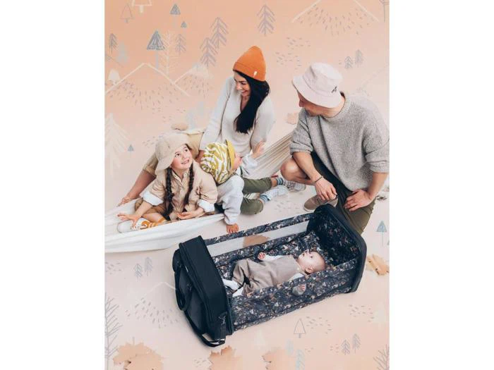 En smilende familie med to voksne og et lille barn sidder på et tæppe, mens en baby sover i en transportabel krybbe på jorden. Baggrunden har et ferskenfarvet pasteldesign med træer.