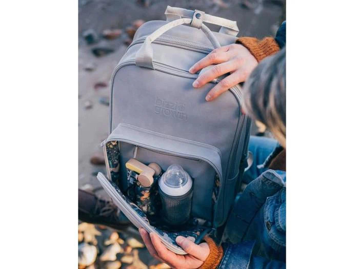 En person åbner en lysegrå rygsæk og afslører en forlomme med en sutteflaske, trælegetøj og andre babyting indeni. Personen er iført en brun sweater og jeans.