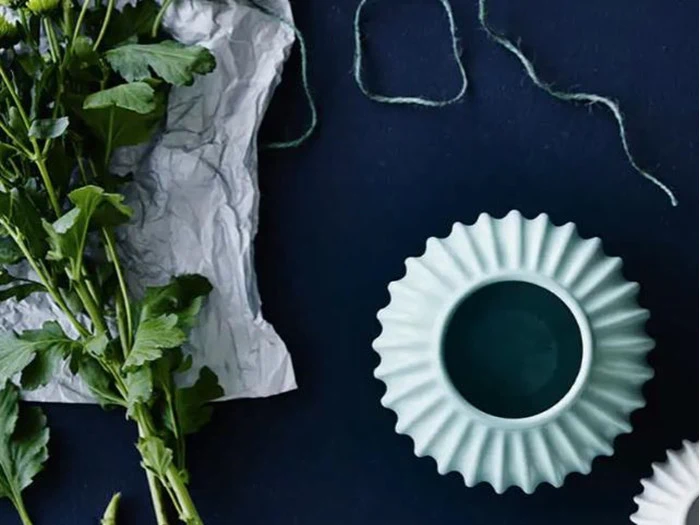 Lysegrøn tekstureret vase ved siden af grønne bladstilke, sammenkrøllet hvidt papir og grønt sejlgarn på en mørkeblå baggrund.