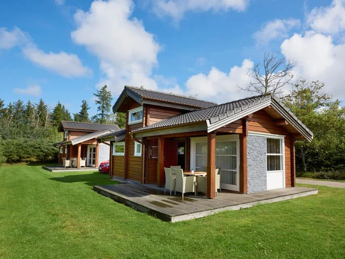 Tre små træhytter med verandaer ligger på en grøn græsplæne, omgivet af træer, under en blå himmel med spredte skyer.