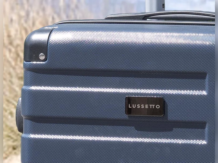 Et nærbillede af en blå kuffert i hård skal med et metallisk LUSSETTO-mærke, udendørs med en sløret naturbaggrund.