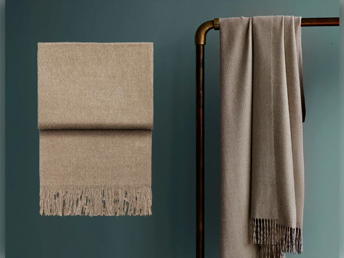 Et beigefarvet tørklæde med frynser er pænt foldet på en væg, og et andet er draperet over et mørkt metalstativ til tøj på en dæmpet grøn baggrund.