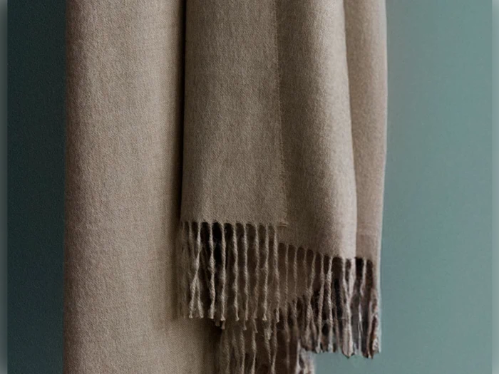 Et beige uldtørklæde med frynsede ender hænger på en dæmpet grøn baggrund.