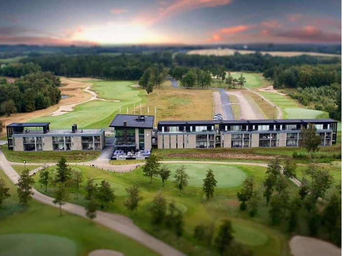 Et moderne hotel med store vinduer ligger ved siden af en golfbane, omgivet af grønne fairways, træer og en dramatisk solnedgangshimmel.