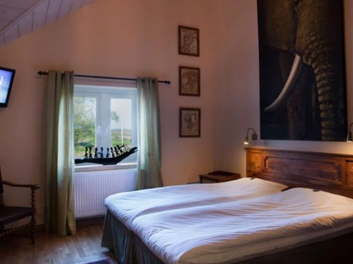 Hyggeligt soveværelse med dobbeltseng, hovedgærde af træ, stort elefantmaleri, vindue med grønne gardiner og et skakspil i vindueskarmen.