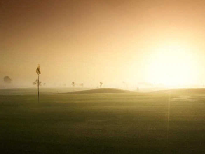 En golfbane ved solopgang med en flagstang i forgrunden, blødt sollys, der kaster et varmt skær over det tågede landskab.