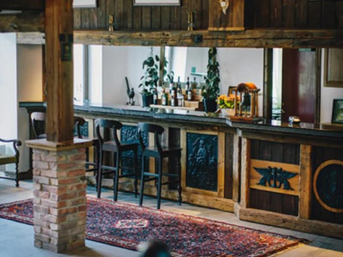 En rustik bar med træstole, en murstenssøjle, potteplanter på disken og et mønstret tæppe på gulvet skaber en hyggelig atmosfære.