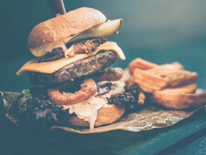 En stablet cheeseburger med salat, løg, ost, en pickles og curly fries, serveret på brunt papir med en side af crinkle-cut fries.