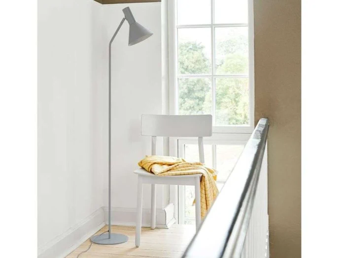 En hvid stol med et gult tæppe lagt over sig står ved siden af en grå gulvlampe ved et vindue, hvor der kommer naturligt lys ind, i et minimalistisk og hyggeligt hjørne.