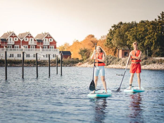 To personer i redningsveste på paddleboard på roligt vand nær en kystlinje med røde og hvide huse og træer i baggrunden.