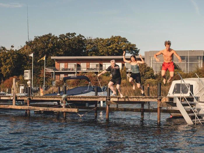 Tre personer i badetøj springer ud fra en træbrygge og ned i en sø med både og bygninger i baggrunden på en solrig dag.