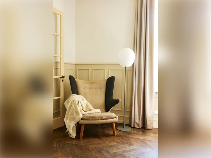 En hyggelig lænestol med et beige tæppe står ved siden af en høj gulvlampe i et lyst, dæmpet rum med trægulv og lyse paneler på væggene.