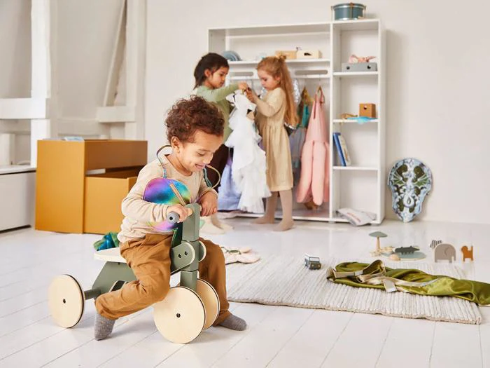 Et lille barn kører på en trehjulet cykel af træ, mens to andre børn leger med udklædningstøj i et lyst og pænt legeværelse.