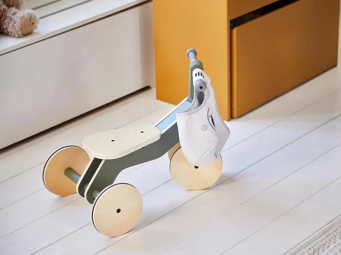 En løbecykel i træ med fire hjul og en lille taske hængende fra styret står på et hvidt gulv ved siden af et gult skab.