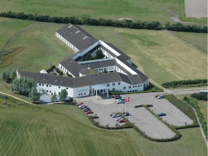 Luftfoto af en stor, hvid, L-formet bygning med en parkeringsplads, tre flagstænger og omkringliggende grønne marker. Flere biler er parkeret foran bygningen.