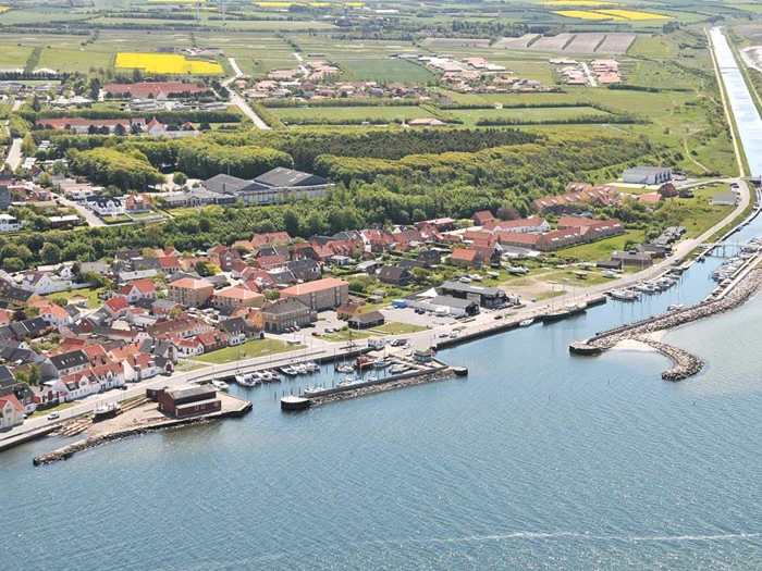 Luftfoto af en kystby med en marina, dokker og både, omgivet af huse, grønne områder og landbrugsjord i baggrunden.