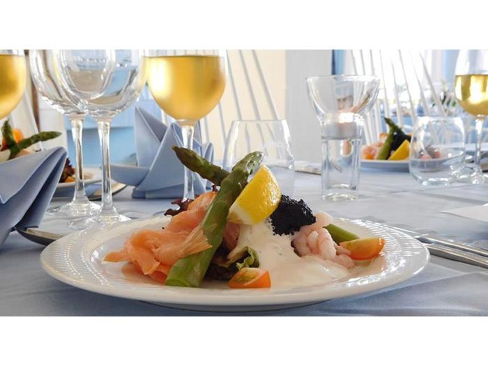 En tallerken med røget laks, rejer, asparges, kaviar, citron og sauce på et dækket bord med vinglas og foldede blå servietter.