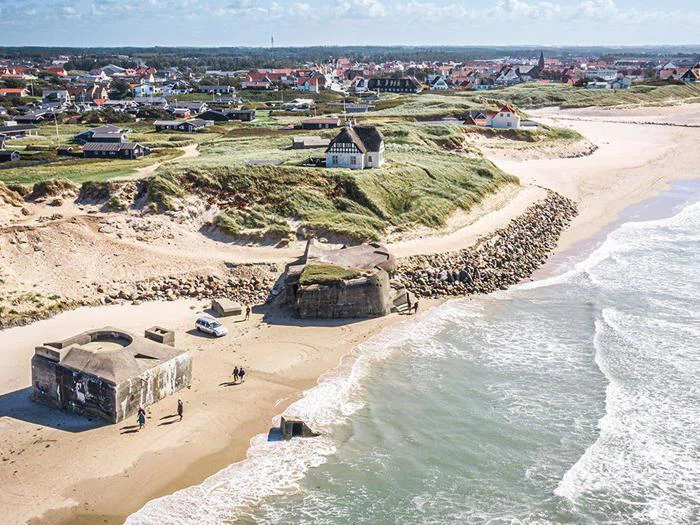 Luftfoto af kystbunkere på en sandstrand med græsklædte bakker, huse og en by i baggrunden under en delvis overskyet himmel.
