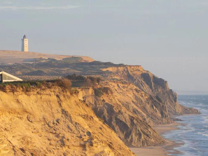 Stejle sandklipper langs en kystlinje med et fyrtårn på toppen, med udsigt over havet under en klar himmel.