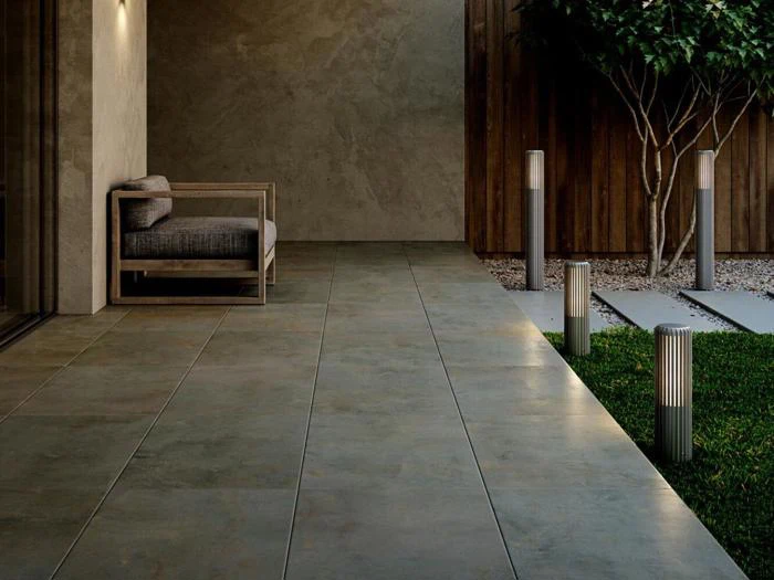 Moderne udendørs terrasse med store grå fliser, en sofa med træramme, dekorativ stibelysning og et træ ved siden af et træhegn.