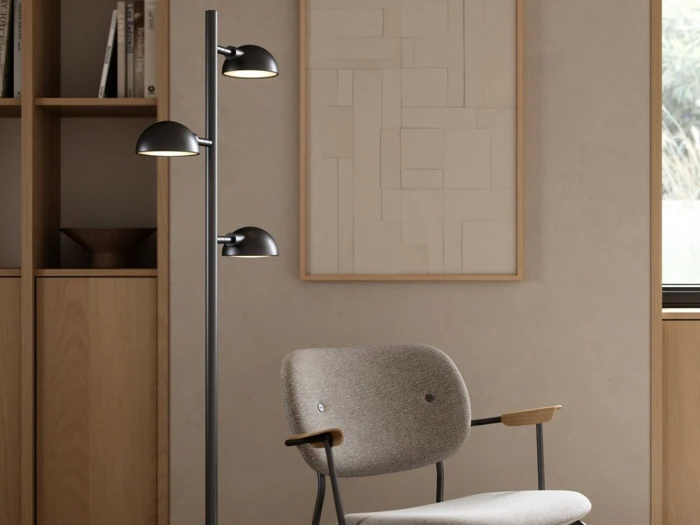 En moderne gulvlampe med to sorte skærme står ved siden af en grå polstret stol i et minimalistisk rum med neutrale vægge, træhylder og et indrammet abstrakt kunstværk.