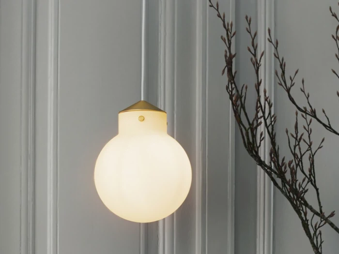 En rund væglampe i matteret glas med guldarmatur lyser blødt på en grå panelvæg ved siden af nøgne trægrene.