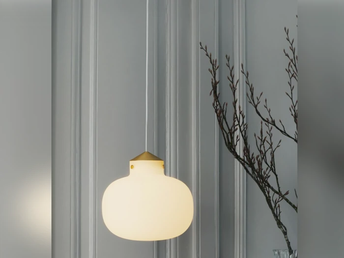 En moderne hvid pendel hænger foran en grå panelvæg med nøgne grene synlige i højre side.