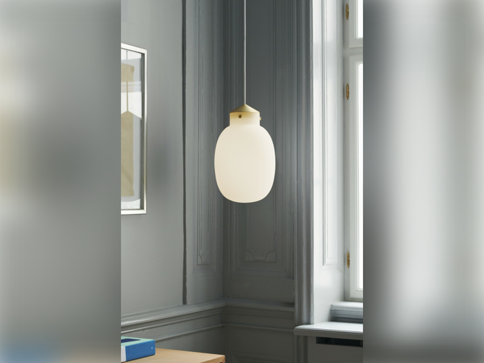 En pendel i matteret glas hænger fra loftet i et gråt rum med dekorative vægstøbninger og et stort vindue, der lukker naturligt lys ind.