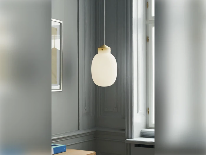 En pendel i matteret glas hænger fra loftet i et gråt rum med dekorative vægstøbninger og et stort vindue, der lukker naturligt lys ind.