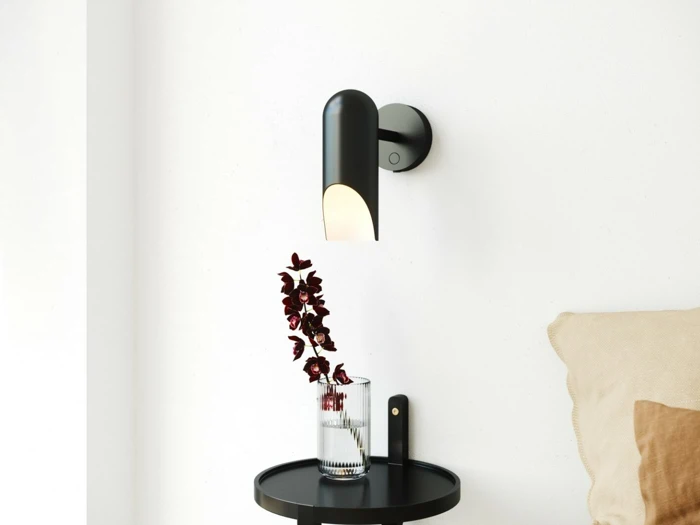 En moderne sort væglampe lyser op på et lille rundt sort bord med en glasvase med mørkerøde blomster ved siden af en beige pude.
