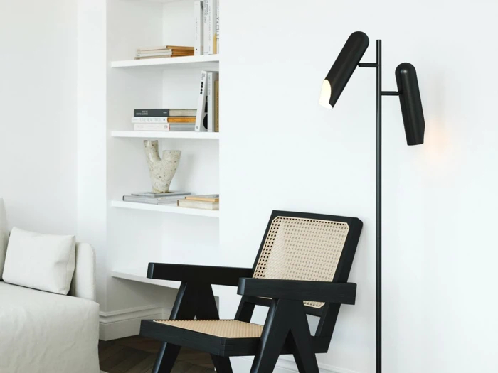 En moderne stue med en sort gulvlampe, en sort stol med vævet sæde, indbyggede hvide hylder og en hvid sofa med puder.