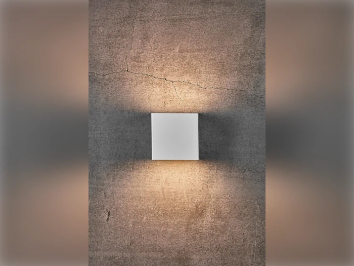 En moderne firkantet væglampe på en struktureret, revnet betonvæg, der kaster varmt lys opad og nedad.