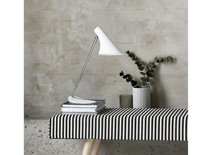 En moderne hvid skrivebordslampe står på en stak bøger ved siden af en stribet bænk og en vase med eukalyptusgrene mod en grå betonvæg.