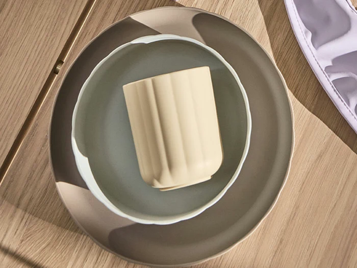 En beige kop placeret i en lysegrå skål, begge anbragt på en større grå og beige tallerken oven på en træoverflade.