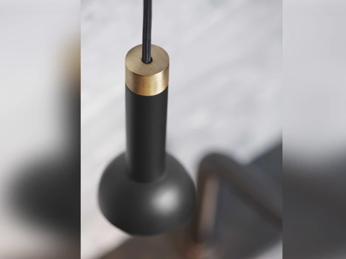 Nærbillede af en moderne sort pendel med en guldmetallisk accent, ophængt i en sort ledning på en sløret, lys baggrund.