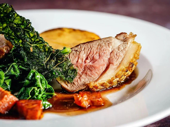 Nærbillede af en anrettet gourmetret med stegt kød i skiver, sprødt grønt, karamelliserede rodfrugter og en rund kartoffel, serveret med en mørk sauce på en hvid tallerken.