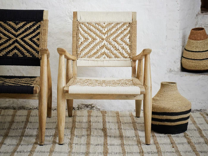 To vævede stole med geometriske mønstre - en sort og en cremefarvet - står på et stribet tæppe ved siden af en vævet kurv mod en hvid struktureret væg.