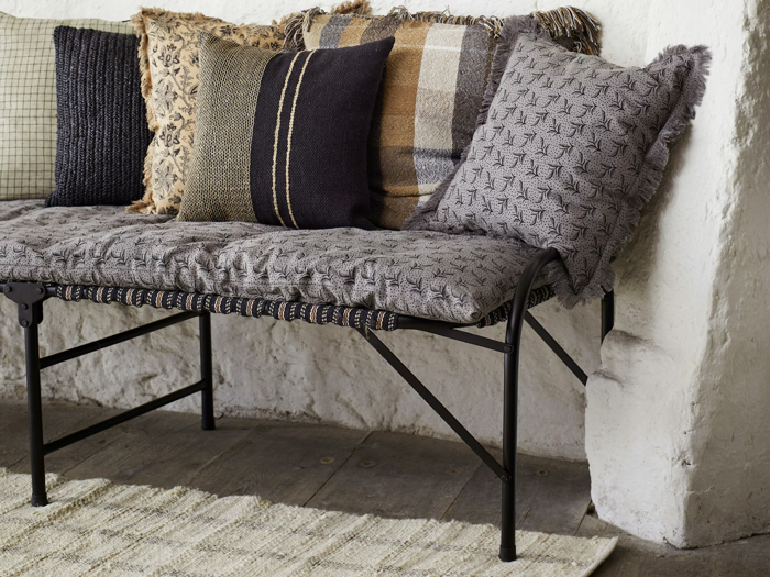 En polstret bænk med mønstrede og ternede puder står mod en hvid struktureret væg på et trægulv.