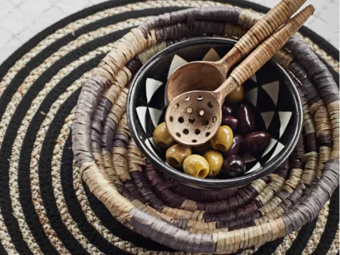 En skål med grønne og sorte oliven med to træredskaber, placeret i en flettet kurv på en sort og beige mønstret måtte.