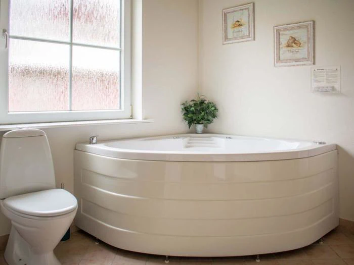 Et rent badeværelse med hjørnebadekar, et hvidt toilet, en potteplante på kanten af badekarret, et stort matteret vindue og to indrammede billeder på væggen.