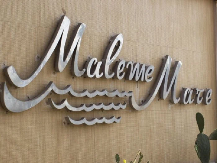 Sølvfarvet metalskilt med teksten Maleme Mare og bølgemotiver på en beigefarvet væg.