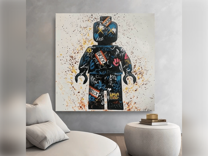 Et stort maleri af en Lego-figur dækket af farverige tags i graffiti-stil hænger på en moderne grå væg over en lys lænestol og en rund ottoman.