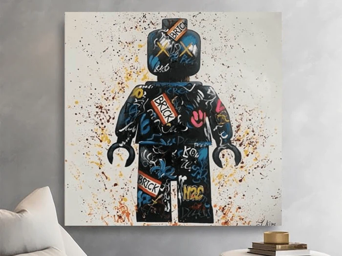 Et farverigt graffiti-maleri af en LEGO minifigur med ord som BRICK og forskellige tags, der hænger på en væg over en sofa og et lille bord med bøger.
