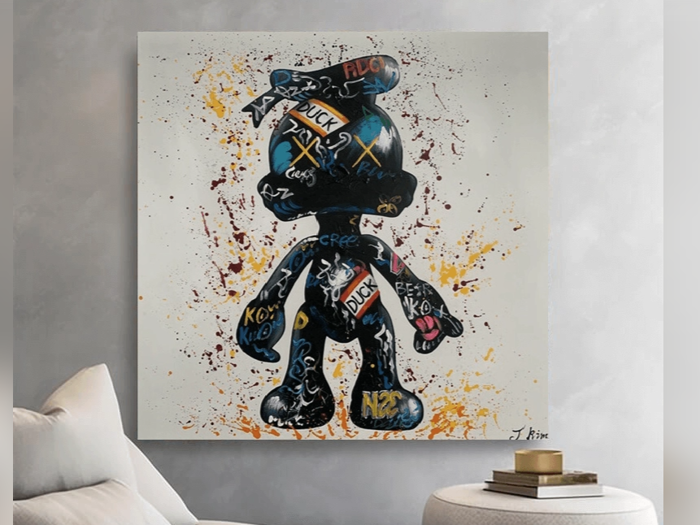 Et moderne maleri af Anders And, dækket af graffitilignende ord og farveklatter, hænger på en lysegrå væg over et rundt sidebord.