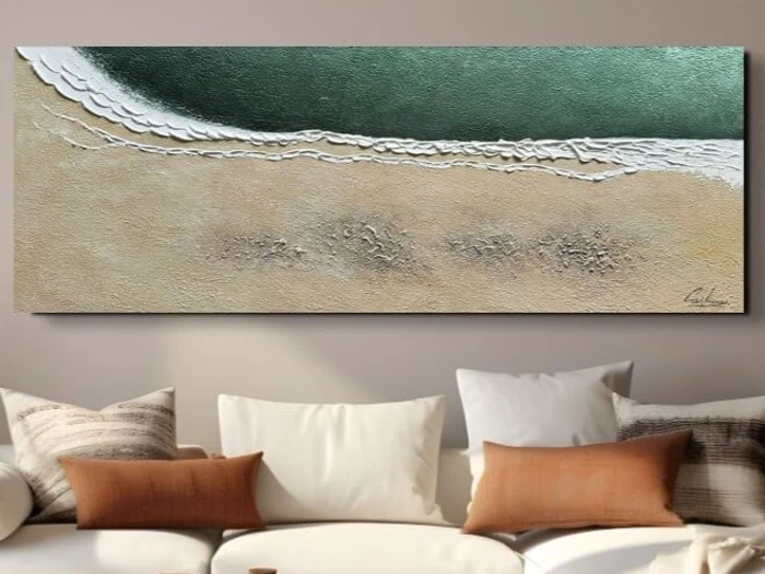Et maleri med struktur, der forestiller havbølger ved en sandstrand, hænger over en hvid sofa med forskellige brune og beige puder.