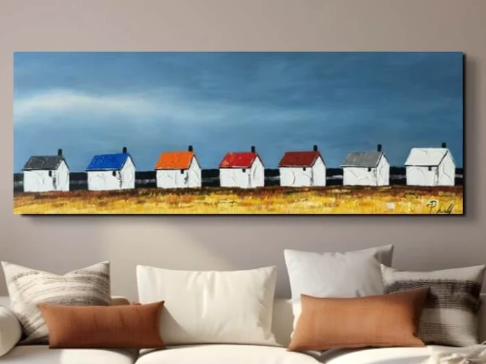 Et maleri over en sofa viser syv hvide huse med tag i forskellige farver, der står mod en blå himmel og en gul mark.