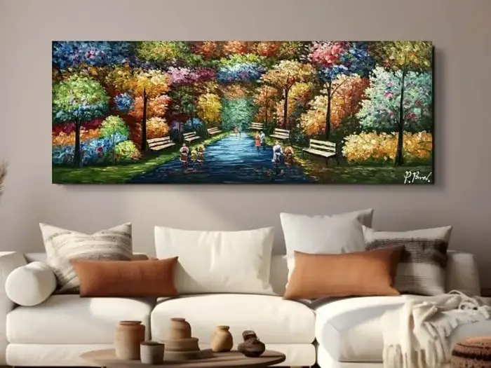 Farverigt landskabsmaleri af en træbelagt sti med bænke, levende løv og gående mennesker, der hænger over en beige sofa med brune og hvide puder i en hyggelig stue.