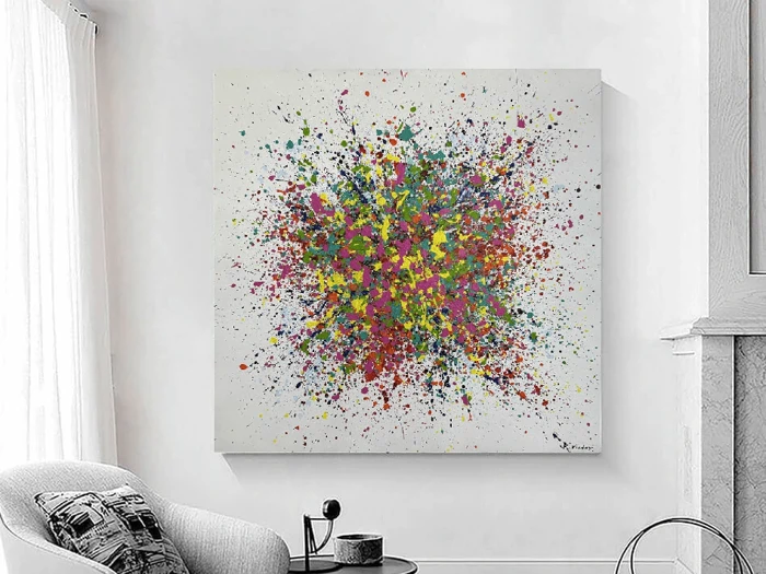 Farvestrålende abstrakt maleri på et hvidt lærred, der hænger på en lysegrå væg i et moderne, lyst rum med en hyggelig stol og et lille bord i nærheden.