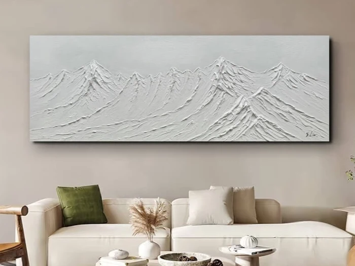 Et hvidt maleri med struktur, der forestiller bjergtoppe på et rektangulært lærred, hænger over en moderne beige sofa med neutrale og grønne puder og minimalistisk indretning.