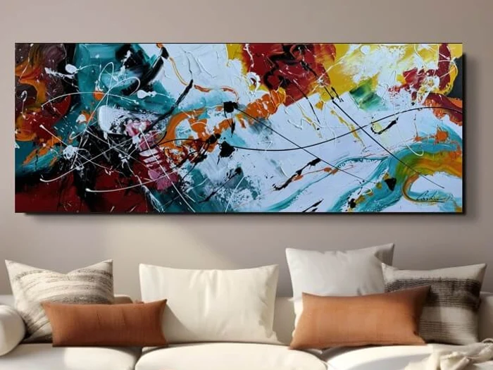 Et stort, farverigt abstrakt maleri med stænk af rød, gul, blå og hvid hænger over en beige sofa med hvide og brune puder.